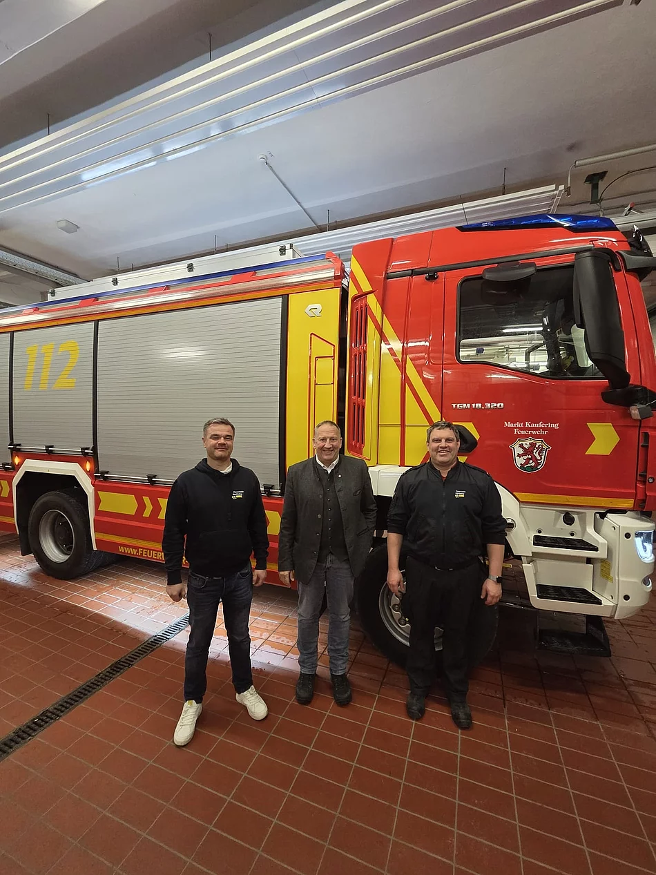 Drei Männer stehen vor einem roten und gelben Feuerwehrfahrzeug mit der Nummer 112 in einer Fahrzeughalle