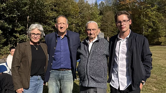 Vier Personen stehen auf einer Wiese vor Bäumen unter blauem Himmel und schauen in die Kamera