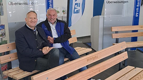 Zwei Männer sitzen auf einer Holzbank in einem Raum mit VR-Bank-Werbefahnen und Informationsständern zu Kontoauszügen und Überweisungen