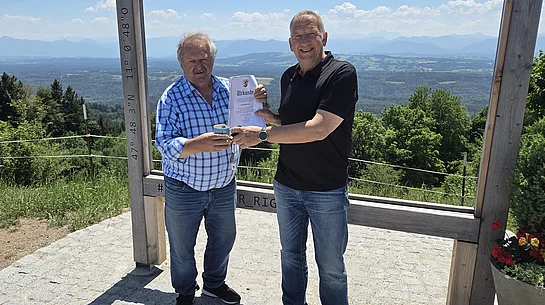Zwei Männer stehen vor einem großen Holzrahmen mit der Aufschrift 'BAYERNS SCHÖNSTER RUNDBLICK', einer hält eine Urkunde, die andere eine Tasse, im Hintergrund Landschaft mit Bergen und blauem Himmel