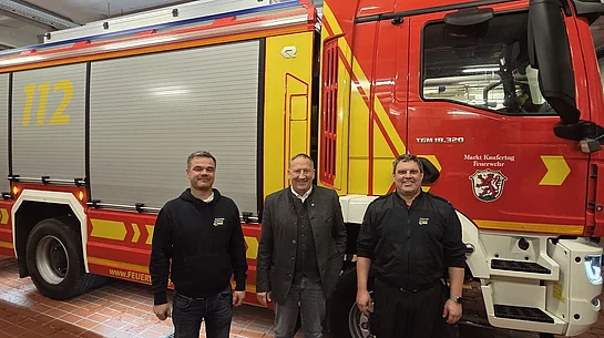 Drei Männer stehen vor einem roten und gelben Feuerwehrfahrzeug mit der Nummer 112 in einer Fahrzeughalle