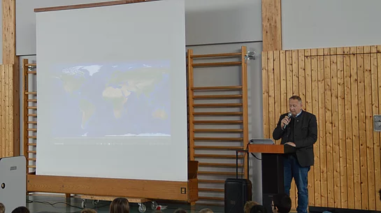 Mann mit Mikrofon steht an Rednerpult vor Leinwand mit Weltkarte, davor sitzende Kinder in Turnhalle mit Holzverkleidung